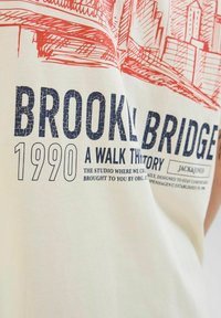 Lys cremefarvet t-shirt med rød grafik af Brooklyn Bridge og marineblå tekst "BROOKLYN BRIDGE 1990". Fremstillet i bomuldsmateriale med blød tekstur.