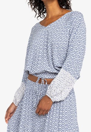 Kobieta ubrana w niebiesko-biały kwiecisty, długi rękaw, krótki top i dopasowaną spódnicę z marszczonym, elastycznym pasem i sznurkiem.