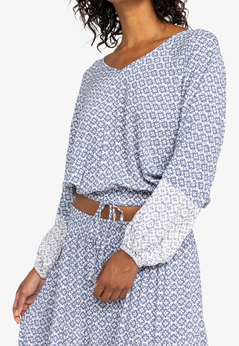 Femme portant un haut court à manches longues à motifs floraux bleu et blanc, assorti à une jupe avec une taille élastiquée froncée et un cordon de serrage.