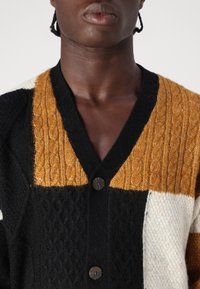 Dziergany kardigan w kolorach czarnym, musztardowym i kremowym; wzory w warkocze; dekolt w serek; dwa ciemne guziki. Teksturowany materiał dodaje wizualnego uroku.