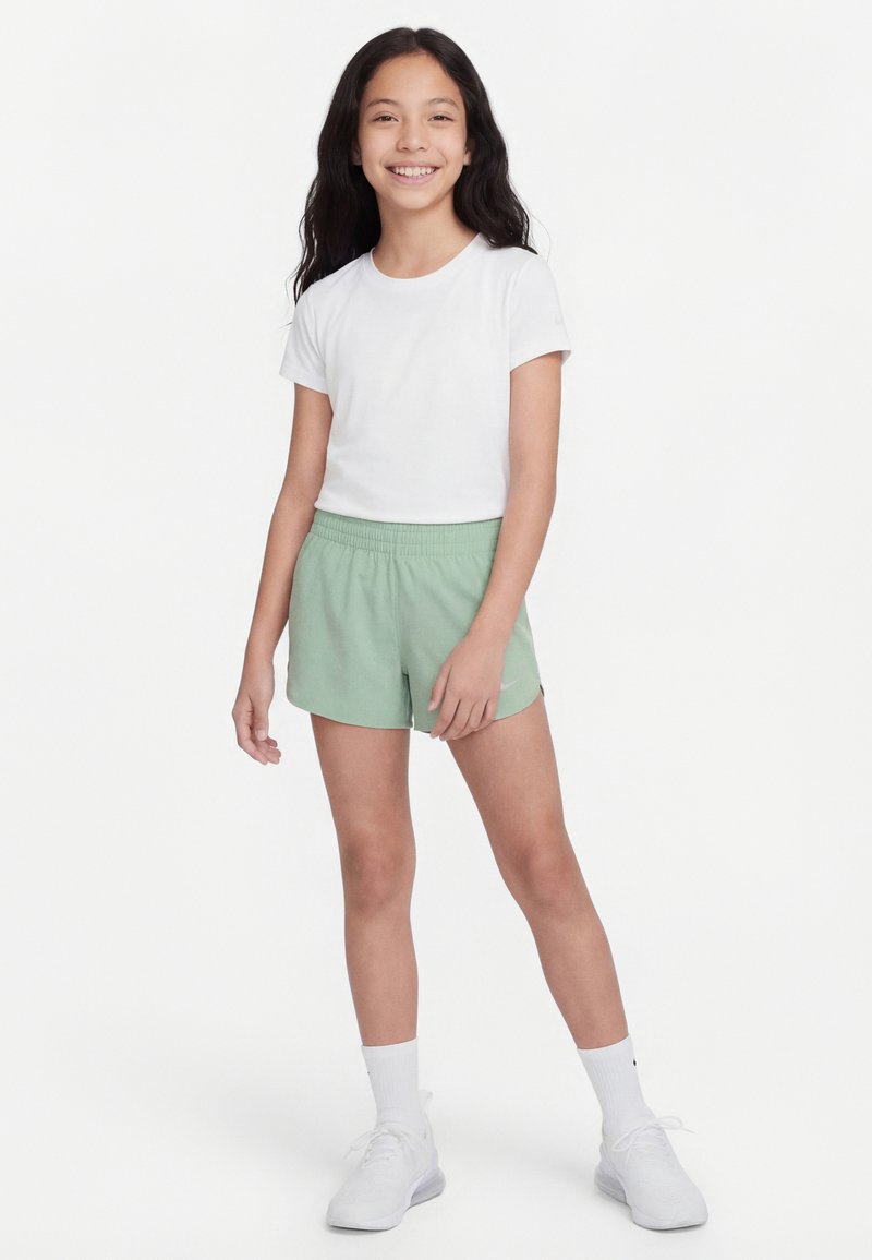 Jeune fille souriante aux cheveux longs et foncés, portant un t-shirt blanc, un short vert clair, des chaussettes blanches et des chaussures de sport blanches, debout.