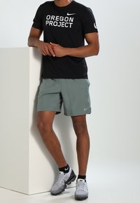 Černé tričko s krátkým rukávem s bílým textem "OREGON PROJECT", v kombinaci se světle zelenými šortkami a šedými sportovními botami s černými akcenty.