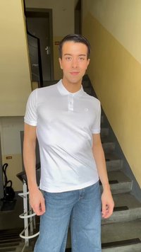 Mladý muž stojaci na medziposchodí schodiska, nosiaci biele polo tričko a modré džínsy, s béžovými stenami a kovovým zábradlím v blízkosti.