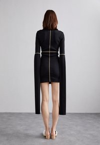 Svart långärmad klänning med guldstickning, figursydd design och förlängda ärmar. Stylad med ljusfärgade blockklackssandaler.