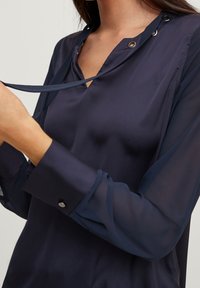 Navy blue long-sleeve blouse made of lightweight, sheer material. Features a split neckline with decorative metallic grommets and buttons at cuffs.