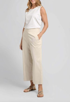 Donna in piedi che indossa una canotta bianca senza maniche, pantaloni beige a gamba larga e corta con tasche, e sandali piatti metallici con cinturini.