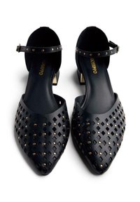 Bailarinas negras con punta afilada, hechas de material texturizado, que presentan un diseño de diamantes calados y tachuelas en tono dorado. Correa ajustable en el tobillo.