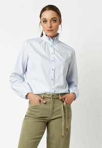 Woman wearing a light blue pinstripe ruffle-neck blouse tucked into olive green high-waisted pants with hands in pockets.