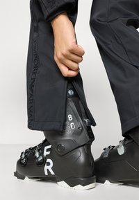 Μαύρο μποτάκι σκι με λευκή επιγραφή "FR" και φερμουάρ σε μαύρο παντελόνι σκι, επιδεικνύοντας σφιχτή εφαρμογή στην περιοχή του αστραγάλου του μποτακιού.