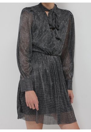 Robe gris foncé pailletée avec manches longues en tulle, nœud noué au col, taille cintrée et jupe plissée portée par une personne debout.