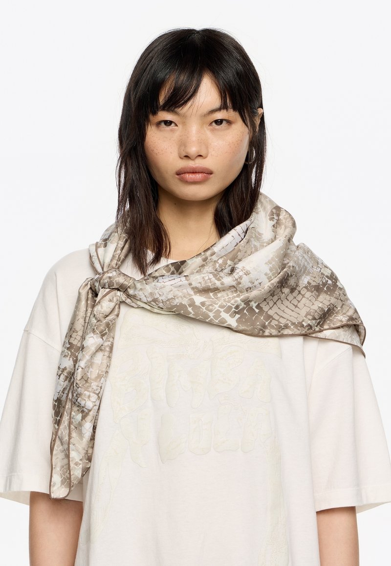 Bufanda ligera con un diseño estampado en tonos beige y crema, con una textura de piel de serpiente, drapeada de forma elegante alrededor del cuello.