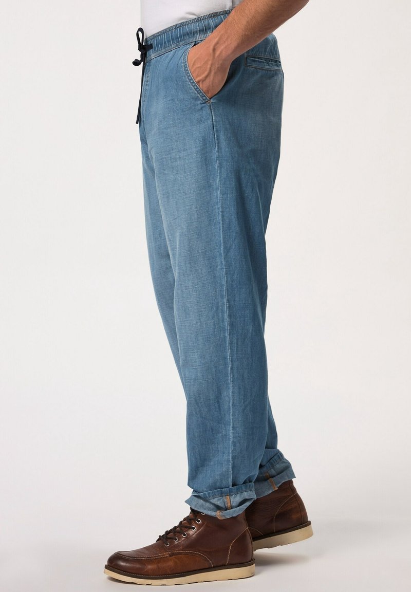 Homme portant un pantalon bleu clair ajusté décontracté avec une taille élastique et des bottes en cuir marron à lacets, debout avec une main dans la poche.