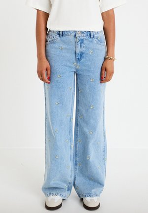 Hellblaue Jeans mit weitem Bein und kleinen gelben bestickten Bienen, kombiniert mit weißen Sneakers und einem weißen Oberteil.
