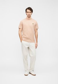 Απαλό ροζ βαμβακερό μπλουζάκι με κοντά μανίκια και μικρό κίτρινο λογότυπο πεταλούδας στο στήθος, συνδυασμένο με μπεζ παντελόνι και λευκά αθλητικά παπούτσια.