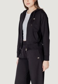 Felpa nera con zip frontale, cappuccio con coulisse e tasche laterali; abbinata a pantaloni della tuta neri coordinati con un piccolo dettaglio del logo.