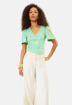 Vrouw met krullend haar die een mintgroene blouse met korte mouwen draagt met een gouden bladmotief en een witte, high-waisted wijde broek.