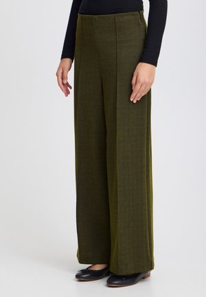 Person trägt olivgrüne, weit geschnittene Hose mit dezentem Fischgrätenmuster, schwarzes Langarmshirt und schwarze Ballerinas und steht.