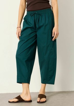 Frau trägt türkisfarbene, kurze weit geschnittene Hose mit Kordelzug in der Taille und braunen Sandalen, steht vor einfarbigem Hintergrund.