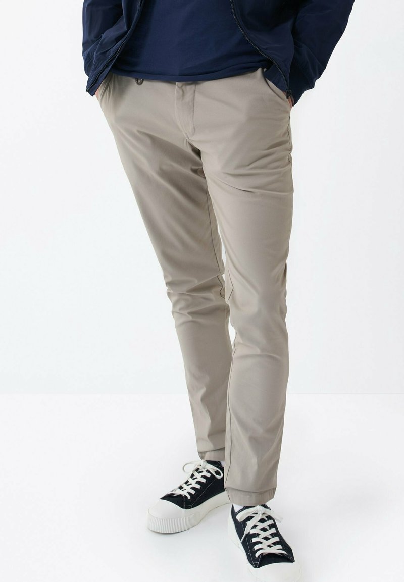 Béžové slim-fit kalhoty s hladkým povrchem a postranními kapsami, spárované s černobílými teniskami se šněrováním.