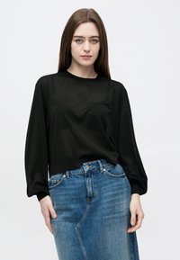 Ung kvinne med langt brunt hår iført en gjennomsiktig svart langermet topp over en BH og et høyt liv blått denimskjørt.