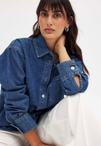 Chaqueta de mezclilla en azul oscuro, con un cierre de botones en la parte delantera, detalles en los bolsillos y mangas enrolladas. El modelo lleva pantalones blancos, de superficie suave.