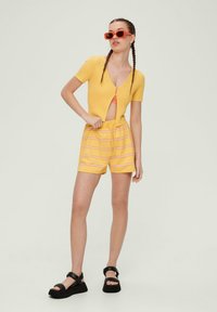 Gelbes geripptes Kurzarmoberteil mit V-Ausschnitt und Reißverschluss vorne, kombiniert mit gelb gestreiften Shorts und schwarzen klobigen Sandalen. Rote Sonnenbrille.