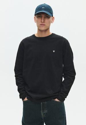 Ung mand iført en navyblå kasket og sort langærmet skjorte med "W.W."-logo, stående med hænderne i lommerne mod en ensfarvet baggrund.