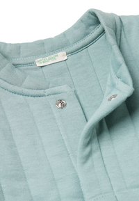 Svetle modrý, žebrovaný pletený oděv s patentkami na límci a štítkem s nápisem "United Colors of Benetton." Měkká textura.