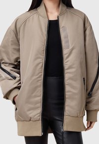 Beige Bomberjacke mit glatter Textur, gerippten Bündchen, Frontreißverschluss und zwei seitlichen Taschen mit Reißverschlüssen. Enthält einfache Designelemente.