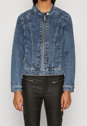Personne portant une veste en denim bleu zippée avec col à boutons-pression et un pantalon noir avec poches zippées.