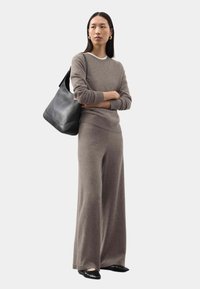 Una donna che indossa un maglione a maglia color taupe e pantaloni a gamba larga, con ballerine nere e orecchini dorati, mentre tiene in mano una grande borsa a tracolla nera, in piedi con le braccia incrociate.