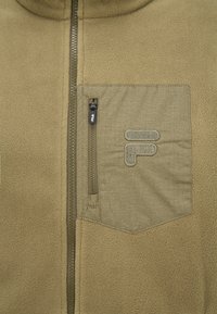 Giacca in pile di colore beige con una texture liscia. Presenta una tasca sul petto con zip e un colletto alto, accentuato da una zip scura e dettagli del logo.