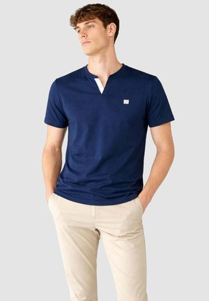 Giovane uomo in piedi con le mani nelle tasche, indossa una camicia a maniche corte blu navy con dettagli di bottoni bianchi e pantaloni beige su uno sfondo semplice.