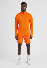 Oranje sportieve zip-up jas en bijpassende shorts, gemaakt van een gladde stof. Bevat witte accenten en een logo op de linkerkant van de borst.
