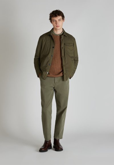 Chaqueta ligera verde oliva con botones, un suéter de punto marrón y pantalones verde claro. Unos botines de cuero marrón completan el look.