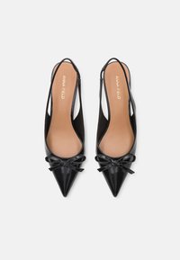 Sorte patentlæder slingback sko med spidse tæer, der har en lille sløjfedetalje foran og en lys indersål med "ANNA FIELD" branding.