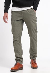 Man som bär olivgröna byxor med sömdekorationer, svart tröja, vit skjorta och bruna läderskor stående med en hand i fickan.