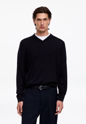 Homme aux cheveux mi-longs et foncés portant un pull noir à col en V sur une chemise blanche, un pantalon foncé et une ceinture noire, debout devant un fond blanc.