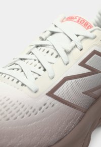 Gros plan sur une chaussure de sport blanche et beige avec une tige en maille, des lacets blancs et un détail de logo près de la semelle.