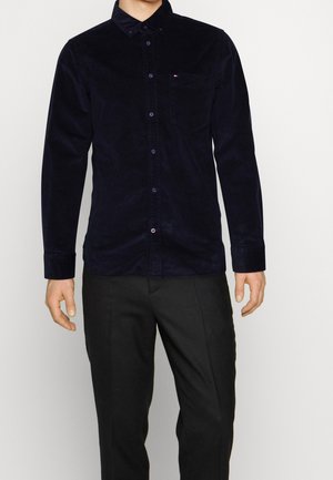 Mand iført en mørkeblå ribbet skjorte med lange ærmer, brystlomme og sorte bukser, der står foran en ensfarvet baggrund.