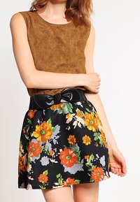 Braunes Wildleder-Top ohne Ärmel, kombiniert mit einem schwarzen Rock mit Blumenmuster, der orangefarbene und gelbe Blumen zeigt, und akzentuiert durch einen schwarzen Schleifen-Gürtel.