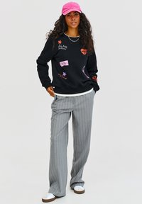 Čierna mikina s farebnými nášivkami, šedé pruhované nohavice a ružová čiapka. Biela košeľa pod ňou a biele tenisky s hnedými podrážkami.