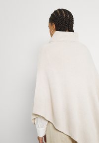 Poncho beige lavorato a maglia con colletto alto e vestibilità ampia, caratterizzato da una finitura testurizzata e un orlo asimmetrico. Materiale morbido.
