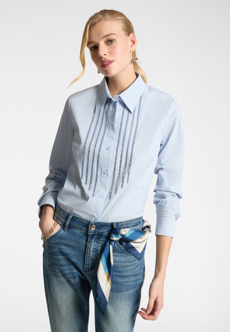 Camicia button-up azzurro chiaro con dettagli a perline verticali sulla parte frontale; maniche lunghe arrotolate ai polsi. Indossata con jeans in denim e una sciarpa a fantasia.