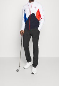 Golfoutfit bestående av en vit, marinblå och röd zip-up tröja, svarta byxor och vita skor, med en golfklubba i höger hand.