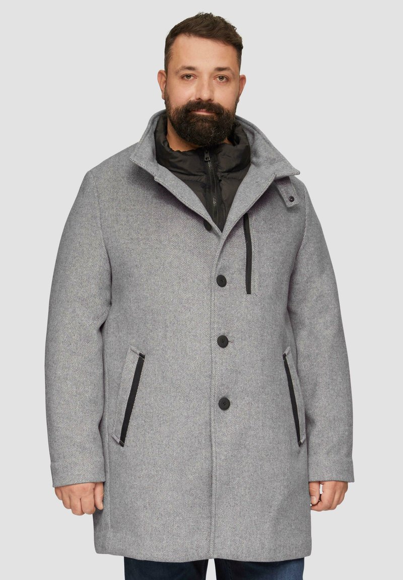 Grijs buitenjas met zwarte accenten, knoopsluiting en een afneembare binnenlaag. Heeft een hoge kraag en zijzakken.