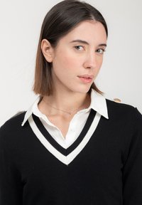 Schwarzer V-Ausschnitt-Pullover mit weißen Streifenakzenten und einem Knopfdetail an der Schulter, getragen über einem weißen Hemd mit Kragen.