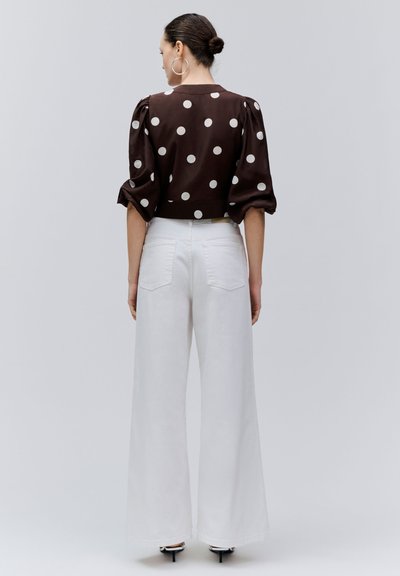 Femme aux cheveux relevés en chignon, portant un chemisier marron à pois avec des manches bouffantes et un pantalon blanc taille haute à jambes larges, debout de dos.