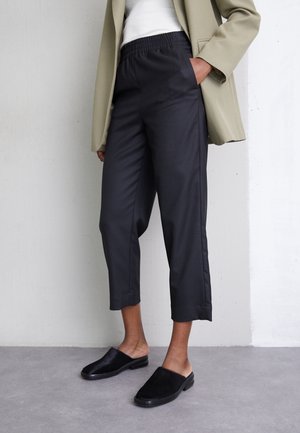 Personne portant un pantalon court noir à taille élastique, un blazer beige, un haut blanc et des chaussures noires sans lacets, debout contre un mur gris.