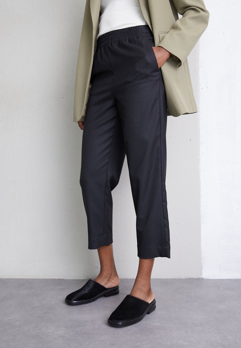 Personne portant un pantalon court noir à taille élastique, un blazer beige, un haut blanc et des chaussures noires sans lacets, debout contre un mur gris.